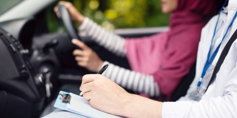 Examiners hand marking a driving test sheet with girl driving in the background